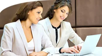 Two business women sitting at a table with a laptop, discussing work projects and sharing ideas.